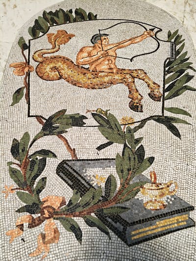 This is a detailed mosaic image depicting a centaur archer drawing a bow and arrow. The centaur has a human torso with muscular arms and a human-like face, and the body of a horse. It is positioned in a leaping or galloping pose, with its rear legs extended and front legs bent. The centaur is looking towards the right of the frame. In the foreground, there are floral elements, including green leaves, brown branches, and orange flowers. There is also a depiction of an open book with a mosaic lamp resting on it, and a cylindrical object that could be interpreted as a scroll or another book. The mosaic is set against a white tiled background.
This mosaic is located on the exterior of the Museo Archeologico Nazionale di Napoli (National Archaeological Museum of Naples) in Naples, Italy. Specifically, it is part of the architectural decoration of the museum's facade.
