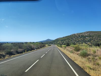 This media file depicts a road trip through a scenic, rural landscape under a clear blue sky, suggesting a sunny day. The perspective is from inside a vehicle, looking forward through a windshield, with the top edge of the windshield and a hint of the car's interior visible in the foreground.
The main subject is a paved road with solid white lines marking the edges and dashed white lines indicating the lanes. The road curves gently to the left as it recedes into the distance. On either side of the road, the terrain is characterized by dry, yellowish grass and scrubby vegetation, with scattered bushes and trees. In the middle ground, a series of low, rolling hills covered in dense green shrubs and scattered rocks rises on the right side of the road. Further in the distance, a conical mountain is visible, also covered in vegetation. The horizon shows a hazy, distant landscape.
The scene appears to be set in a warm, dry climate, typical of rural Spain, specifically Valencia de Alcántara, Spain. There are no people or animals visible in the image. The overall mood is one of peaceful travel and exploration of natural beauty. The time of day is clearly daytime, likely late morning or early afternoon, given the bright sunlight and clear sky. There are no discernible text elements within the image itself.