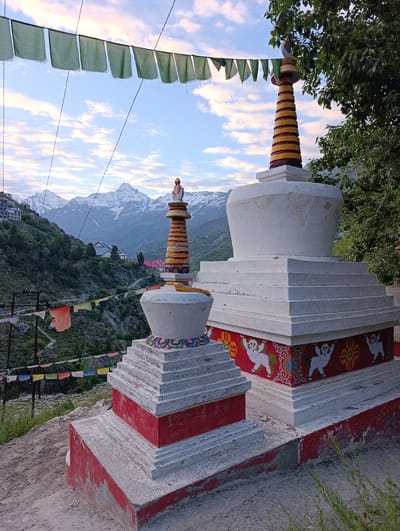 In Kardang, India, two Buddhist stupas stand prominently on a hillside overlooking a valley with snow-capped mountains in the distance. Prayer flags, a mix of green and orange, are strung across the scene, adding a touch of color against the pale blue sky scattered with soft clouds. The stupas are constructed with white, tiered bases, the larger one featuring decorative red and white motifs of mythical creatures. Both stupas are topped with colorful, spire-like structures. The surrounding landscape is a mix of rugged, green terrain with sparse trees and some buildings visible on the slopes. The lighting suggests either early morning or late afternoon, with a soft, diffused light illuminating the scene.