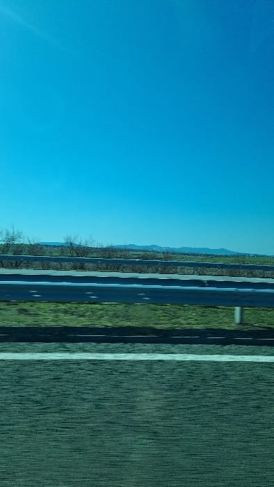 The scene depicts a rural landscape in Krumovo Gradishte, Bulgaria, viewed from a vehicle on a road under a clear sky. In the foreground, a dark asphalt road surface is visible, featuring a solid white lane marking positioned horizontally. Immediately beyond the road, a silver-colored metal guardrail stretches across the mid-ground. Behind the guardrail, a strip of green grass extends, sparsely populated with leafless deciduous shrubs and small trees. In the distant background, a series of low hills or mountains are discernible on the horizon. The upper two-thirds of the image are dominated by a clear, vibrant blue sky, with a bright glare in the upper left quadrant indicating the presence of a strong light source, likely the sun. No individuals or specific actions are observed.