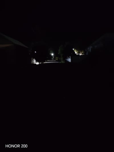 The image is a dark, nighttime scene, likely taken in the city of Kakaydi, Uzbekistan, as indicated by the watermark "HONOR 200" which appears to be a device identifier and not a location. The view is of a street with buildings on either side. A faint light source is visible in the distance, possibly a streetlamp, illuminating a portion of the road. The overall impression is one of low visibility due to the darkness, with the structures and environment being largely obscured. No people or specific actions are discernible.