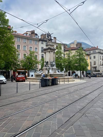 The image depicts a street scene in Grenoble, France, centered around the Fontaine des Trois Ordres on Place Notre-Dame. Tram tracks run along the paved road, curving towards the right of the frame. The fountain is surrounded by buildings of varying heights, with apartments and shops visible. A mountain is visible in the background. People are standing near the fountain, some interacting with it. Vehicles, including a white sedan and a red car, are parked along the street. Trash bins are positioned near the fountain. Overhead tram wires crisscross the sky. The weather appears overcast.