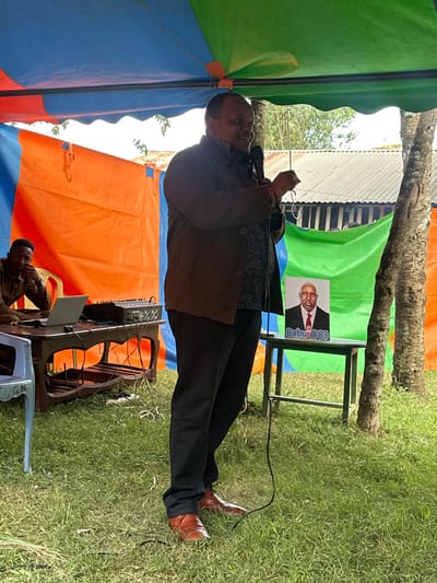 The image captures an outdoor event taking place in Kiganjo, Kenya, under temporary shelters constructed from colorful tarpaulins (green, blue, and orange). The main subject is a man standing and speaking into a microphone. He is dressed in a dark brown jacket over a patterned shirt, black trousers, and brown pointed shoes. He appears to be bald and is actively engaged in addressing an audience not visible in the frame.

Behind the speaker, to his right, is a banner displayed on a stand. The banner features a smiling portrait of an older man in a suit and red tie. Prominently visible text on the banner reads "Babu@83", suggesting a celebration or commemoration, possibly a birthday, for someone named Babu who is 83 years old. There also appears to be additional, smaller text or a handle below "Babu@83".

To the speaker's left, another individual is seated on a white plastic chair, operating electronic equipment. A laptop is open on a small white table, adjacent to what appears to be an audio mixer or sound control board, with various cables connected. This person is wearing a brown collared shirt.

The setting is a grassy area with large tree trunks visible in the background, reinforcing the outdoor nature of the event. The bright lighting indicates it is daytime. The overall scene suggests a public address, a community gathering, or a celebratory function, organized with temporary infrastructure.