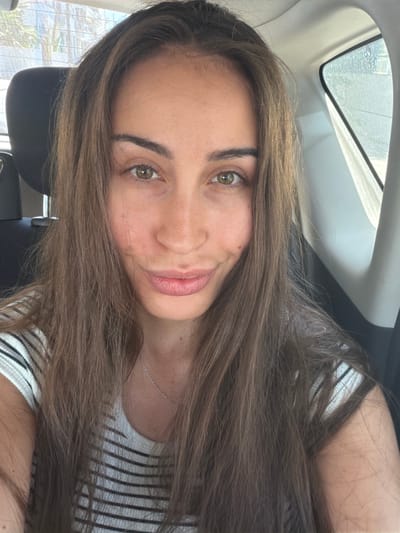 A medium shot selfie of a young woman with long, brown hair, taken inside a vehicle. She is looking directly at the camera with a slight smile. Her eyes are hazel, and she has light skin with a few freckles visible on her cheeks and nose. She is wearing a black and white striped t-shirt and a delicate silver necklace. The background shows the interior of a car, including a headrest, window with a partial view of the outdoors which appears to be a city with palm trees and buildings, suggesting the location is Larnaca, Cyprus.