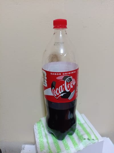 A Coca-Cola bottle with a red cap stands on a white and green striped cloth. The bottle is mostly full of dark liquid and has a label that says "SABOR ORIGINAL" in white text with a FIFA World Cup logo. The Coca-Cola logo is in white script on a red background, and there is a soccer ball design on the label. The bottle appears to be made of clear plastic. In the background, a lightly textured wall is visible. There is text printed on the bottle near the cap that reads "L51005144BPF VAL 10/11/26". The scene is likely indoors, given the lighting and wall texture. There are no people or animals present. The overall impression is that of a product shot or a simple depiction of the beverage. The location context of Saurimo, Angola, does not provide any direct visual cues within the image itself.
