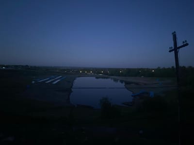 The image depicts an outdoor scene at dusk in Южный, Russia. A body of water, likely a lake or large pond, is centrally located with dark, still water reflecting the sky. The shoreline is visible, showing a beach-like area with some light-colored rectangular structures that could be tents or shelters. In the background, there's a line of trees and a distant town or city illuminated by streetlights. To the right, a wooden utility pole stands prominently, casting a shadow. The foreground is mostly dark, obscuring details, but some foliage is visible.