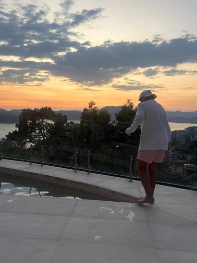 An elevated view from a poolside terrace in Valle de Bravo, Mexico, depicts a person observed from behind during sunset. The sky displays an orange and yellow gradient at the horizon, transitioning to light blue and scattered gray clouds overhead. Multiple layers of dark hills and mountains are visible in the distance, framing a large body of water, likely the lake characteristic of Valle de Bravo. Dense green trees occupy the midground slopes, partially obscuring a distant urban area with scattered structures visible on the right.
In the foreground, a light-colored paved terrace extends across the frame. To the left, an infinity-edge pool with water is partially visible. A transparent glass railing with several stainless steel posts runs along the edge of the terrace, overlooking the landscape.
A single individual, a human, stands on the paved terrace, facing away from the camera and looking towards the distant view. The person is barefoot, with distinct wet footprints visible on the light-colored tiles. They are dressed in a white long-sleeved top or light jacket, pink and white striped shorts, and a white baseball cap featuring a dark emblem on the rear. The individual's right leg is slightly lifted, indicating a motion of walking or shifting stance. No other individuals are present within the frame.