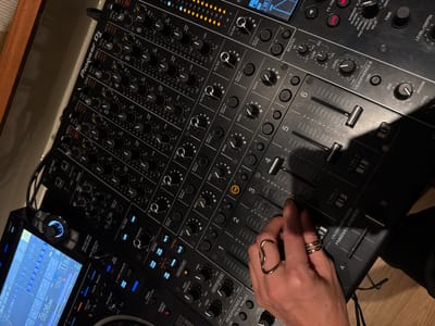 The image shows a close-up, high-angle view of a Pioneer DJ mixer. The mixer is black with various knobs, sliders, and buttons. Several channels are visible, with numbered labels from 1 to 6. The screen of a connected DJ controller or media player is partially visible on the left, displaying waveform data and tempo information. A person's hand with rings is interacting with the mixer, touching a slider. The environment appears to be indoors, with warm-toned wooden flooring visible at the bottom right. The lighting is dim, with strong highlights and shadows, suggesting artificial lighting. There are no visible indicators of the city of Unknown, Maldives in the image.