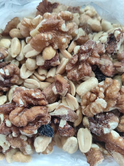 The image displays a close-up, overhead view of a mix of nuts, primarily walnuts and peanuts, with a few dark raisins scattered throughout. The nuts are contained within a translucent plastic bag, the top edge of which is visible in the upper portion of the frame. The nuts appear to be lightly coated with a powdery substance, possibly sugar or salt. The lighting is bright, casting subtle shadows that highlight the textures of the nuts. There is no discernible information about the location in Askiz, Russia, within the image itself.