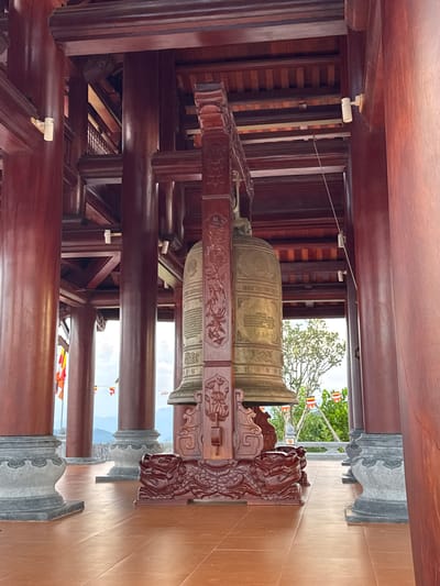 In the city of Diên Khánh, Vietnam, a large, ornate bell hangs within a wooden structure. The bell is made of bronze and features intricate carvings. It is supported by a wooden stand decorated with dragon motifs. The structure is supported by thick, dark red wooden pillars and beams. In the background, colorful prayer flags are visible, as well as lush greenery and distant mountains under a cloudy sky. The ground is paved with reddish-brown tiles.