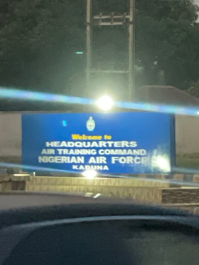 The image shows a sign that reads "Welcome to HEADQUARTERS AIR TRAINING COMMAND NIGERIAN AIR FORCE KADUNA". The sign is illuminated by a light source. The scene appears to be at the location of the Headquarters Air Training Command of the Nigerian Air Force in Kaduna. The surrounding environment appears to be a gate or entrance area with foliage visible in the background. The location is likely within the city of Kaduna, near Hayin Banki.
