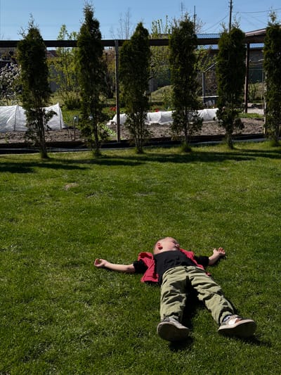 A boy lies on a lawn. He wears green pants, a black shirt, and a red overshirt, along with sneakers. The lawn is bordered by trees and a fence, possibly in a backyard. A white greenhouse is visible through the trees. The image appears to be taken in daylight. The location is Kurylivka, Ukraine.