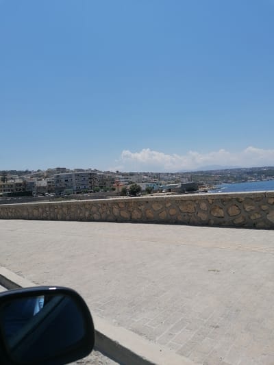 The image shows a view of the city of Rethymno, Greece, from a car. In the foreground, a car's side mirror is visible on the left, reflecting a blue object. The ground is paved with light-colored bricks. Behind the pavement, a stone wall runs horizontally across the frame. Beyond the wall, the sea is visible on the right, with a city skyline on the left and in the distance. The city features numerous buildings of various sizes and architectural styles, with some structures appearing to be hotels or apartment complexes. The sky is clear and blue, with a few scattered white clouds.