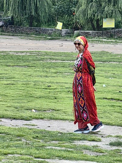 A woman dressed in traditional attire, including a red and yellow headscarf, sunglasses, a red embroidered tunic, and dark pants with athletic shoes, is walking on a dirt path in a grassy area. She is wearing large silver earrings and a necklace. The background features a stone wall, trees with long, drooping branches, and two yellow signs. The setting appears to be a park or open space, potentially in Solang, India, given the context provided, with lush green grass and a dry, dusty path. The woman is looking to her left, and there are no other people visible in the immediate vicinity.