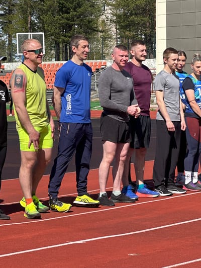 A ground-level, outdoor, color photograph shows a group of seven people standing in a line on a red running track in Polarnie Zori, Russia. The track has white lane markers. The people are all looking to the right of the frame.

From left to right: The first person wears neon green shorts and a light green shirt, sunglasses, and neon green and orange socks with neon green shoes. There is a tattoo on the person's left arm. The next person wears a blue t-shirt with white lettering and navy blue pants with yellow sneakers. The next person has on a gray long-sleeved shirt and black shorts with black sneakers. The next person wears a burgundy t-shirt and black shorts with blue sneakers. The next person has on a gray long-sleeved shirt and black pants with black sneakers. The last two people wear blue shirts with black pants and black and white athletic shoes.

Behind the people are a building and trees. A basketball hoop is visible behind the first person. The lighting is bright.