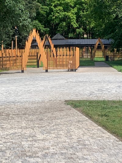 The image depicts an outdoor park area with a paved pathway in the foreground and a wooden fence structure with a covered pavilion in the background. The pathway is made of small, grey interlocking stones. Beyond the pathway, there is a green lawn area. The fence is constructed from vertical wooden planks arranged in a decorative, arched pattern. Behind the fence, a dark-colored pavilion with a sloped roof is visible. The environment is lush with mature green trees surrounding the area. There are no visible people or animals in the image. The scene appears to be in a well-maintained public space, possibly a park or recreational area.