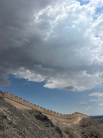 The image shows a section of the Great Wall of China, specifically a part of the Jiayuguan Pass, located in Jiayuguan, China. The wall is constructed from brick and appears to be ancient, with battlements along its crest. It is situated on a rocky, arid hillside, with sparse vegetation visible. The sky above is a mix of dramatic, dark storm clouds and brighter, partly cloudy blue sky. The lighting suggests it is daytime, possibly with the sun breaking through the clouds. In the distance, a flat, developed landscape can be seen, indicating the presence of a city or settlement. There are no people or animals visible in the frame. The perspective is from a lower vantage point, looking up at the wall as it snakes along the undulating terrain.