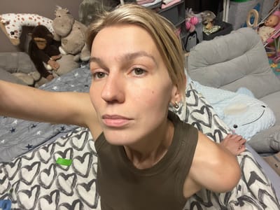 A young woman with light blonde hair and fair skin is wearing a brown tank top and lying on her back on a bed with a light gray and white comforter with a pattern of black hearts. The comforter has a piece of green tape on it. She is looking directly at the camera with a neutral expression. Her right arm is outstretched towards the camera and her left arm is bent with her hand resting on the comforter. The bed is in a room in Moscow, Russia (latitude: 55.659, longitude: 37.591). The room appears to be a child’s room with a stuffed animal giraffe, hippopotamus, and monkey visible on the bed behind her. There is a large grey armchair with light grey pillows in the background. A stuffed penguin toy is visible on the right side of the image. An open shelf with clothes is visible in the background behind the woman’s head. There is a baby stroller and a baby doll sitting in it in the upper right corner of the image. The scene is a bedroom, possibly in a family home in the city of Moscow. The lighting in the room is well-lit. The photo was taken from a low angle, as if the camera is being held by the woman herself. The woman appears to be posing for the photo.