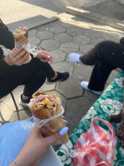 The image shows an outdoor scene with three people sitting or standing on a gray, hexagon-patterned pavement. Two of the people are holding wrapped sandwiches or shawarma-like foods in their hands.

One person, wearing black leggings with white dots and white sneakers, is partially visible in the upper left corner. Another person is wearing black pants and black sneakers. The third person, positioned to the right, is wearing light blue jeans. The hand holding the sandwich has blue nail polish and a silver bracelet.

A blanket with a green and white floral pattern, along with a red and white checkered bag, is visible in the upper right corner.

The location is Satovcha, Bulgaria.