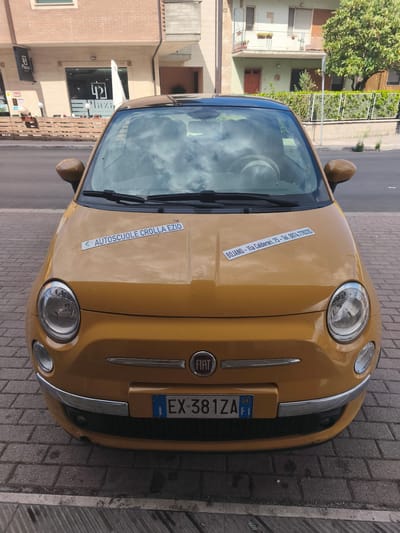 This is a medium-shot, eye-level photograph of a yellow Fiat 500 parked on a brick sidewalk. The car is facing the viewer directly. The front of the car has two round headlights and a chrome grille with the Fiat logo in the center. A license plate is visible on the lower front of the car, reading "EX-381ZA".
There are two rectangular stickers on the hood of the car. The left sticker reads "AUTOSCUOLE CROLLA EZIO" in blue text. The right sticker is longer and reads "BOJANO - Via Calderari, 75 - Tel. 0874.778335" in blue text.
In the background, a building with a brick facade and several windows is visible. There is a sign on the building that says "Plaza" with a logo. To the right of the building, there is a green tree and a fence. The scene appears to be outdoors during the daytime. The sky is reflected in the windshield of the car, showing clouds. The weather appears to be sunny. The location is indicated by the text on the sticker as Bojano, Italy, with the street address Via Calderari.