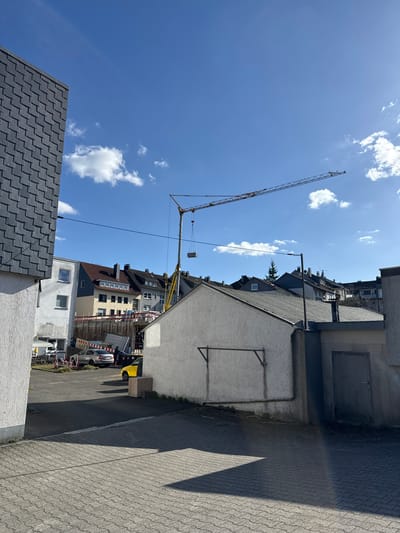 The image shows an urban scene in Remscheid, Germany. A tall yellow crane dominates the sky, positioned over a construction site nestled amongst older buildings. The construction site is bordered by a road and a sidewalk paved with grey bricks. Adjacent to the sidewalk is a building with a white, slightly weathered exterior. Part of a dark grey slate roof is visible in the lower left corner. The sky is blue with scattered white clouds. The buildings surrounding the site appear to be a mix of residential and commercial structures. There is a yellow dumpster along the side of the street.