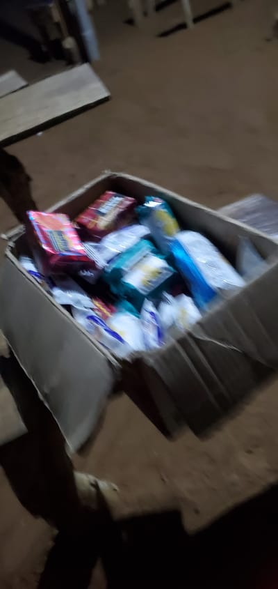 A cardboard box filled with assorted packaged goods, primarily food items like biscuits or snacks, sits on the ground. The items inside are of various colors, including red, blue, green, and white packaging. The box appears to be damaged or worn. The surrounding environment suggests an outdoor setting, possibly a dirt or unpaved ground with some wooden planks or debris visible in the background. The lighting is dim, indicating it might be evening or an indoor area with low light. The image quality is poor, with motion blur affecting many of the details. The location is identified as Numan, Nigeria. There are no visible people clearly depicted in the scene, only dark, blurred shapes that could be shadows or partially obscured figures.