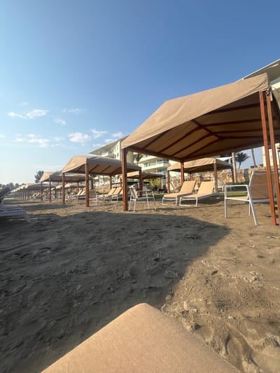 The image depicts a beach scene, possibly in Acapulco de Juárez, Mexico, given the presence of beach umbrellas and lounge chairs arranged in rows along the sand. The sky is clear with some scattered clouds. The beach umbrellas are uniform in design with brown canvas tops and brown metal frames. The lounge chairs, appearing to be made of a similar material to the umbrellas, are placed under the umbrellas. The sand is of a dark color and shows the footprints of beachgoers. In the foreground, part of a lounge chair is visible. The photograph is taken at an angle, which makes it appear that the image is on its side.