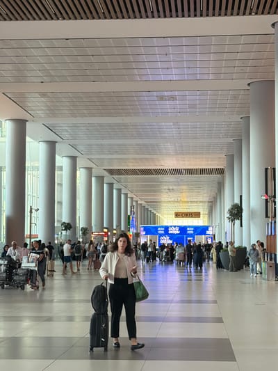 The image depicts a large, modern airport concourse in Arnavutköy, Turkey. A woman is walking towards the camera in the foreground, pulling a black suitcase and carrying a green tote bag. She is wearing a white jacket, a light-colored top, and black pants. Her right hand is holding a mobile phone. The concourse features a high ceiling with a distinctive slatted wooden upper section and a grid of illuminated panels below. Numerous large, cylindrical white pillars line the walkway, creating a sense of depth and scale. In the mid-ground, other travelers are visible, some with luggage, and a row of shops and service counters can be seen. A yellow directional sign with Turkish text "← ÇIKIŞ" (Exit) is prominently displayed. The floor is tiled with a pattern of grey and white squares. The overall atmosphere is that of a busy, functional transportation hub.