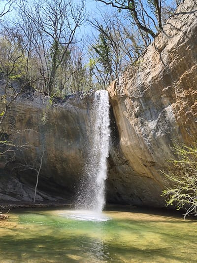 A waterfall cascades into a clear, green-tinted pool at the base of a rocky cliff, flanked by trees. The scene is located in the Орлиновский округ, Ukraine.