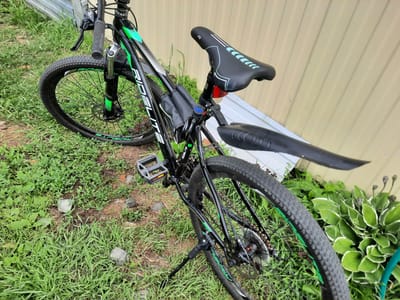 The image shows a black RIDE LITE bicycle parked on a grassy area next to a corrugated metal fence. The bicycle has green accents on the frame and wheels, a black saddle with teal accents, and a black kickstand. A black saddlebag is attached to the frame under the seat. The foreground is dominated by lush green grass and some rocks, while the background consists of the beige corrugated metal fence and some green foliage. The scene appears to be outdoors, likely in a residential area, given the presence of the fence and the cultivated plants. The lighting suggests it is daytime. There are no people or animals visible in the image. The location is specified as Bol'shoye Sorokino, Russia.