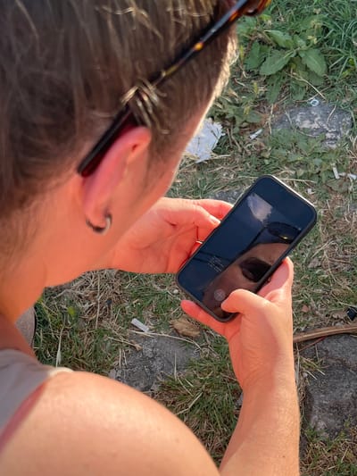 The image shows a person's head and hands holding a smartphone. The person has short, light brown hair and is wearing sunglasses. The smartphone screen reflects the person's face and the surrounding environment. The person is outdoors, with grass and rocks visible in the background. The setting appears to be a park or a natural area. The overall lighting suggests it is daytime, possibly late afternoon due to the warm tones. There is no specific indication of the location being Podostrog, Montenegro.