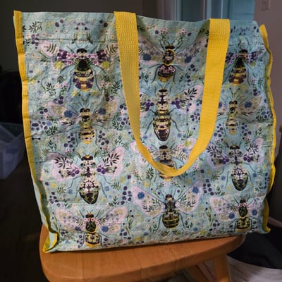 The media file shows a close-up, overhead view of a quilted tote bag with a light blue background and a repeating pattern of stylized bees surrounded by small flowers and leaves. The bees are depicted with black and yellow stripes on their abdomens, and their bodies are filled with floral designs in shades of white, purple, and green. Two yellow fabric straps are attached to the bag, one running across the top and the other diagonally over the patterned fabric. The bag appears to be made of cotton or a similar fabric, with visible stitching lines that indicate its quilted construction. The edges of the bag are trimmed with a bright yellow fabric. The bag is resting on a light brown wooden surface, likely a chair or table, with some darker background elements suggesting an indoor setting. The lighting is ambient and evenly distributed. No people, text, or specific time of day or weather cues are visible. The overall impression is of a handmade item with a charming, nature-inspired design.