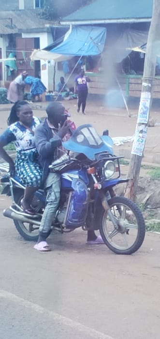 The scene depicts a bustling outdoor market area in Mua ward, Kenya, with a blue motorcycle and its two occupants prominent in the foreground.
**Foreground:** A dark-skinned male, wearing a dark jacket, light cargo pants, and light pink slides, is seated as the driver of a blue motorcycle equipped with a light blue windshield. He is seen holding a dark object, possibly a phone, to his right ear and has dark sunglasses on. Behind him, a dark-skinned female passenger is seated, wearing a blue and white patterned dress and dark sandals, with her hands resting on her hips. The motorcycle is positioned on an unpaved or dirt road. To the immediate right of the motorcycle is a wooden utility pole adorned with several flyers or posters, one clearly displaying "25" in white text on a blue background.
**Midground/Background:** The area behind the motorcycle reveals an active market environment. Several individuals are present: one woman in a light-colored top and skirt is bending over, seemingly inspecting goods on the ground. Another person, possibly a woman, in a blue garment, is also visible near market stalls. Further back, a person in a purple t-shirt with the number "25" printed on it is walking from left to right. The market consists of several makeshift stalls utilizing blue and white tarpaulins for shelter, supported by poles. Various goods, possibly produce or packaged items, are visible on the ground. Buildings with corrugated metal roofs and light-colored walls line the background, indicating permanent structures alongside the temporary market setups. The overall lighting suggests daytime conditions.