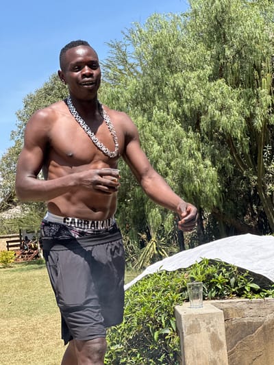 An outdoor scene in Olkaria ward, Kenya, depicts a shirtless Black male standing centrally under a clear blue sky. He wears a large, silver-colored chain necklace, black boxer briefs with "CARRRALKALAN" visible on the waistband, and black shorts. He holds a clear plastic cup containing a dark beverage in his right hand. His left arm is extended forward with a partially clenched fist. His gaze is directed towards the viewer. The background features dense green foliage, including tall trees with light green leaves and a cactus-like plant. A white tarp or sheet partially covers a low green bush in the mid-ground. A clear drinking glass rests on a concrete block in the lower right. In the distant left background, two small, indistinct figures are visible seated on a wooden structure within a grassy area. The scene is brightly illuminated by direct sunlight.