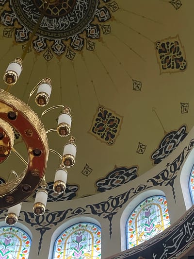 The image shows the interior of the Al-Azhar Grand Mosque in Bekasi, Indonesia, focusing on the dome's interior and a large chandelier. The dome is painted with intricate geometric and calligraphic patterns in muted colors. Several lamps hang from the chandelier, which is made of gold-colored metal. Below the dome are arched stained-glass windows with floral motifs. The overall impression is one of ornate and decorative architecture within a place of worship.