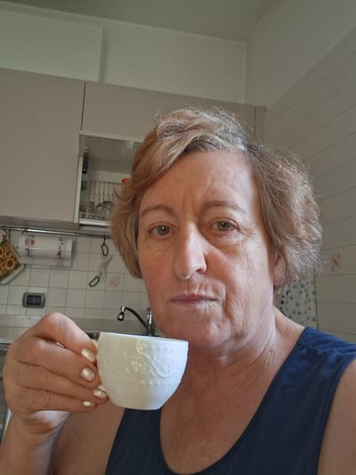 The image shows a woman holding a white coffee cup. She is in a kitchen with white tiled walls and cabinets. A faucet is visible behind her. The woman has short, blonde hair and is wearing a blue tank top. Her nails are painted a pearly white color. The scene appears to be a candid photograph taken indoors. The location is identified as Padua, Italy.