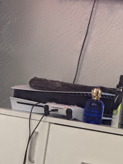 The image shows a PlayStation 5 game console stored inside a white cabinet or shelving unit. On top of the console are several items, including a bottle of cologne. A black cable is connected to the console and extends towards the right side of the frame. The console is positioned near a gray wall. It's not possible to determine the location in Glojene, Bulgaria from the image alone.