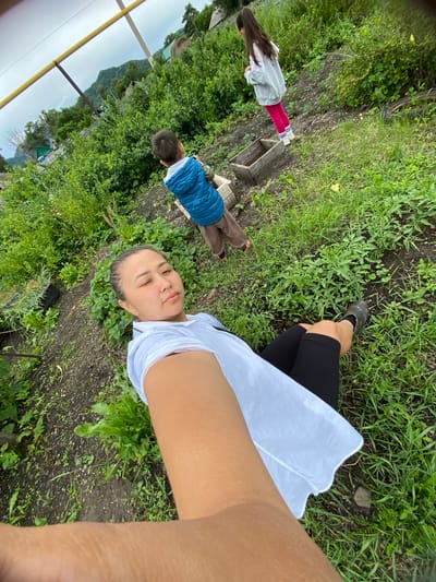 In Maloarslangulovo, Russia, a woman is lying on the ground in a garden, holding a camera to take a selfie. She is wearing a white shirt and black leggings. A young boy in a blue vest and brown pants is standing to her left, and a young girl in a gray hoodie and pink pants is standing to the boy's left. The garden is filled with green plants and dirt. In the background, there are houses and trees, with mountains in the distance. The sky is overcast.