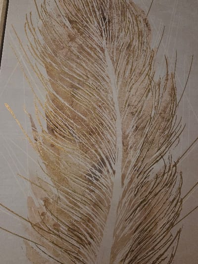 This image displays a close-up view of a vertically oriented artwork depicting a stylized feather. The dominant color palette consists of various shades of brown, beige, cream, and white, extensively highlighted with metallic gold accents. A prominent, broad central rachis, creamy-white in color, extends vertically through the feather. Barbs emanate from this rachis, rendered in a combination of crisp white lines, textured brown watercolor-like washes, and distinct metallic gold linear details and flecks that suggest a reflective quality. The background is a uniform light beige or cream, featuring subtle, abstract geometric lines and soft brown smudges that integrate with the feather's overall design. The artwork appears to be a painting or print on a canvas or textured surface. No individuals, specific actions, or additional environmental elements are present within the depicted artwork. The scene is located in Blainville-sur-Orne, France.