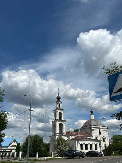 The image depicts a street view in Yerino, Russia, dominated by a bright blue sky with large, fluffy white clouds. In the center of the frame, a white church with a bell tower stands prominently. The church has a domed roof with a reddish-brown patina and a bell tower topped with a black spire and a cross. Adjacent to this church, to the left, is another building with a bright blue roof and a cross.
In the foreground, two dark-colored cars are parked on the side of a paved road. A street lamp with two lights is visible between the church and a line of trees. A blue and white road sign indicating a speed bump is positioned on the right side of the image, with branches of foliage encroaching on it. The scene is framed by green trees on the left and right edges, with a low fence in front of the buildings. The overall atmosphere is that of a sunny day in a semi-rural or suburban area.
