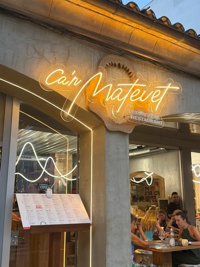 The image shows the exterior of a restaurant called "Ca'n Matevet Vermuteria Restaurant" located in Port D'alcúdia, Spain. A neon sign in bright yellow spelling out the restaurant name is prominently displayed above the entrance. Below the sign, a clear plastic sign indicates "VERMUTERIA RESTAURANT". To the left of the entrance, a menu is displayed on an easel inside the restaurant, visible through the glass. A sign reading "OPEN" is also visible near the menu.
Inside the restaurant, patrons are seated at tables, some of whom are interacting and dining. In the foreground, a group of women are seated at a table, engaged in conversation and enjoying their meal. One woman on the right is wearing a green top and has a glass of red wine. Another woman to her left is wearing a blue top and is also seated at the table. Further back, a man in a black t-shirt is standing and looking towards the patrons. The interior appears to be furnished with bookshelves and other decorative elements. The overall ambiance suggests a lively dining establishment.
