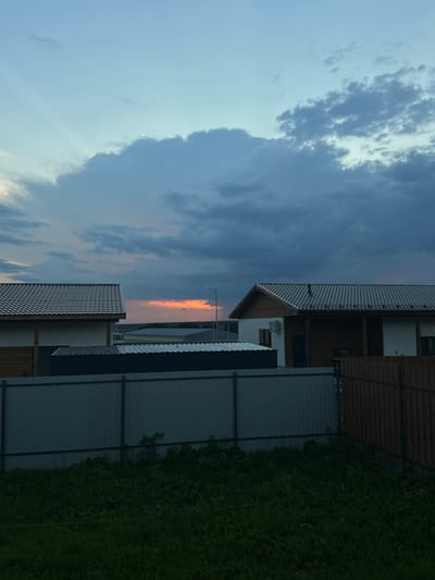 The image depicts a suburban landscape at dusk, likely in Rassokha, Russia. Two houses with dark, shingled roofs are visible, one on the left and one on the right. Both structures feature wooden and white siding. A dark metal fence runs horizontally across the foreground, separating the houses from a patch of dense, green grass. In the distance, between the two houses and beyond the fence, a strip of the sky glows with vibrant orange and yellow hues, indicating a sunset. Dark, heavy clouds dominate the upper portion of the sky, contrasting with the brighter sunset colors. A streetlamp is visible on the right side of the image, near the house. The overall scene is one of quietude, with no people or animals present.