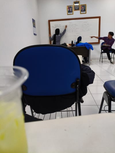 The scene depicts an indoor classroom or meeting room in Muka, Indonesia. A woman, dressed in a gray long-sleeved shirt and black hijab, stands facing a large, wood-framed whiteboard on the rear wall, actively writing on it with her right hand. Above the whiteboard are three framed portraits, including a central emblem flanked by two male figures in suits. To the woman's right, a man wearing a patterned long-sleeved shirt sits on a chair, pointing his right index finger towards the whiteboard or the woman, with an open notebook in his left hand. A dark brown desk with two dark water bottles sits in front of the whiteboard. The room features white walls and a white tiled floor with dark grout lines. In the immediate foreground, out of focus, is the blue backrest of a chair with a dark garment draped over it, and in the extreme left foreground, a blurry translucent cup containing a yellowish liquid rests on a white surface.