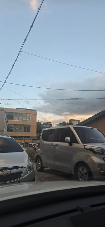 The image shows a street scene in Gimhae-si, South Korea, under a cloudy sky with hints of sunset colors. Two silver cars are parked on a paved surface. The car in the foreground on the left appears to be a Chevrolet Spark, with its front grille and headlights visible. To the right, a larger silver van, possibly a Kia Carnival, is parked. A tan building with multiple windows is visible behind the cars, and further back, other buildings and structures can be seen. Electrical wires crisscross the sky above the scene. A person is partially visible sitting on a low wall or curb behind the van, wearing light-colored clothing. The scene depicts an ordinary urban environment during dusk.