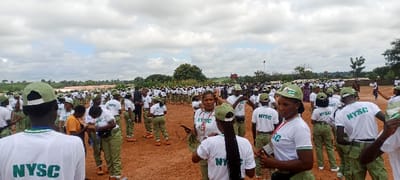 The image captures a large outdoor gathering of approximately several hundred individuals on an unpaved, reddish-brown dirt field in Peyer, Nigeria. The sky is predominantly overcast with patches of brighter cloud. The majority of subjects are observed wearing a uniform consisting of white t-shirts with "NYSC" printed in green, olive-green caps featuring a Nigerian flag emblem, and olive-green trousers. Many individuals wear lanyards with identification cards around their necks. Subjects in the foreground are standing and engaged in casual interactions; one female subject in uniform, positioned centrally, faces the camera with a slight smile, holding sunglasses and a cap in her left hand. Another female subject to her right also looks towards the camera. The crowd extends densely into the background, where distant green foliage, scattered trees, and indistinct low-rise structures are visible.