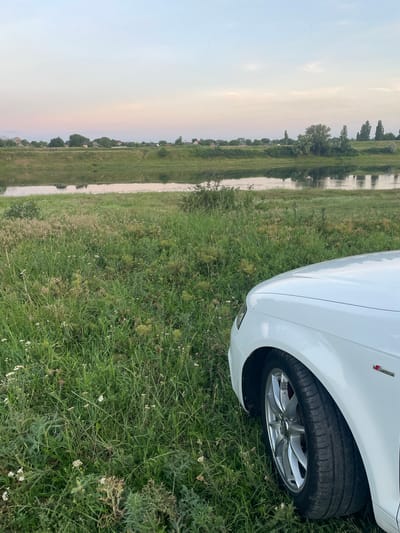 This image, taken in Coșnița, Moldova, depicts a portion of a white car, specifically its front fender and wheel, parked on a grassy verge. The car is angled, with its front right wheel clearly visible, showcasing a silver alloy rim. Beyond the car, a long, narrow body of water runs parallel to a field of tall green grass dotted with small white flowers. The water reflects the sky and the trees lining its opposite bank.

The landscape suggests a rural or semi-rural setting, with hints of buildings visible amongst the trees on the far side of the water. The sky is a pale, hazy blue transitioning to soft pink and orange hues near the horizon, indicating either dawn or dusk. The overall lighting is diffused, suggesting an overcast or hazy day, or the twilight hours. There are no people visible in the frame.

A small red and silver emblem, likely a car manufacturer's badge, is visible on the car's fender. No other text is discernible in the image. The scene evokes a sense of tranquility and a connection with nature, with the car positioned as an observer of the serene landscape.
