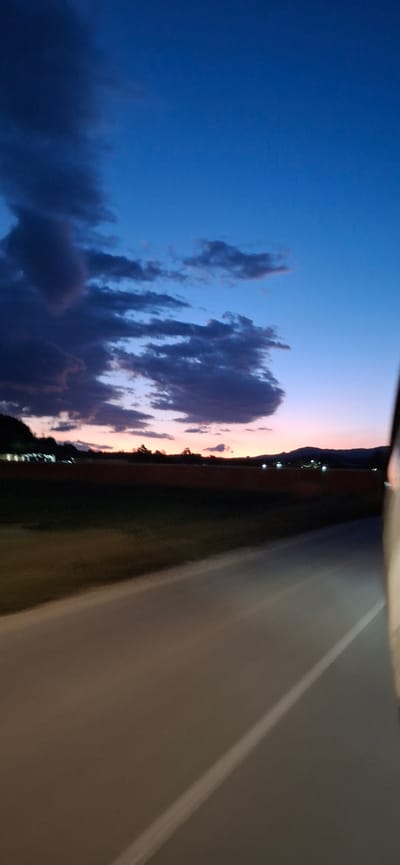 The image depicts a twilight scene with a deep blue sky transitioning to a soft pink and orange hue near the horizon. Dark, dramatic clouds are scattered across the upper portion of the sky. Below the sky, a dark silhouette of hills and distant lights can be seen. In the foreground, a paved road runs diagonally from the lower right corner towards the center, with a faint white line marking its edge. The road appears to be slightly blurred, suggesting movement, possibly from a vehicle. To the left of the road, a strip of dark land is visible, possibly a field or open area. The overall impression is of a rural landscape at dusk, potentially on the outskirts of Ablanitsa, Bulgaria, given the context. No people or specific actions are discernible, but the scene suggests a journey or observation from a moving vehicle.