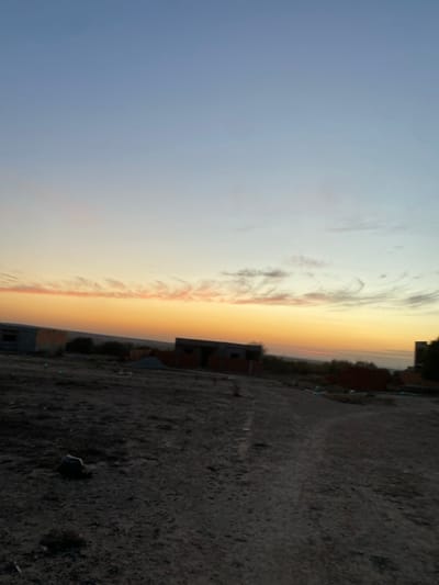 A wide-angle shot captures a landscape divided between sky and land. The sky is a gradient of blue to orange, with textured clouds. Below, the land is a dark, uneven surface, possibly dirt or gravel. Scattered brick structures suggest construction or abandoned buildings. The scene exudes a sense of solitude and desolation, heightened by the perspective and color contrast.