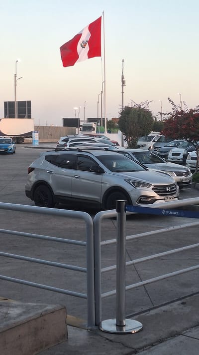 The image captures an outdoor scene, likely at a transportation hub or parking area, with the Peruvian flag prominently displayed on a flagpole in the upper left portion. The flag is red and white with a crest in the center. In the foreground, a silver SUV with black rims is parked, partially obscured by a metal railing system. Behind the SUV, several other vehicles, including sedans and vans, are parked in rows. A white truck with a trailer is visible in the background. The environment appears to be an urban setting, possibly near the border crossing into Chile, given the presence of the Peruvian flag and the overall infrastructure. The sky is a pale orange, suggesting either dawn or dusk. The ground surface is asphalt or concrete. There are no people clearly visible in the image. The context strongly suggests the scene is located in Tacna, Peru, a city known for its proximity to the Chilean border and its importance as a transportation and commercial center.