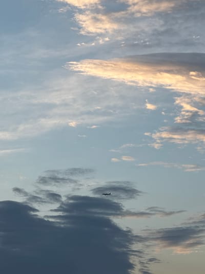 A small airplane is seen flying in a pale blue sky with scattered clouds. Some of the clouds are tinged with orange and yellow, suggesting it might be sunrise or sunset. The lower portion of the image shows darker, more dense clouds. The scene is likely a view of the sky above Misaylovo, Russia.